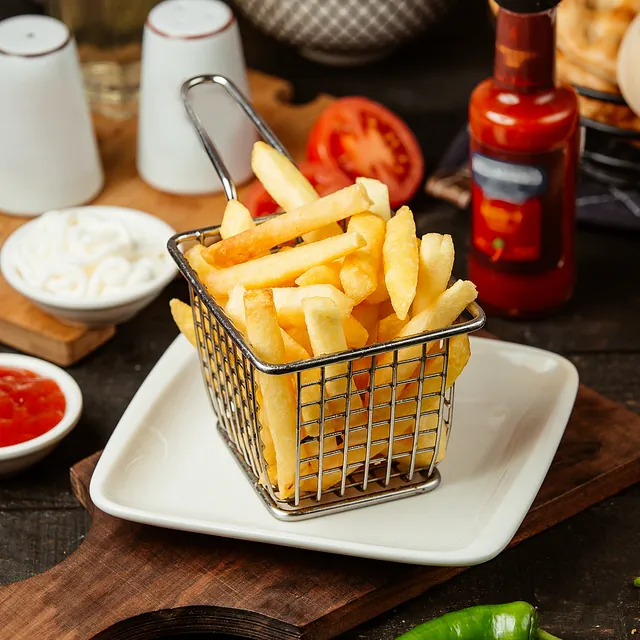 Pommes frites in einem kleinen Metallkorb auf einem weißen Teller mit Ketchup und Mayonnaise im Hintergrund
