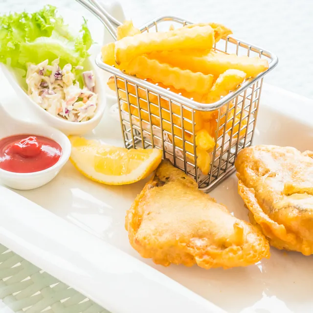 Pommes frites in einem kleinen Metallkorb auf einem weißen Teller mit frittiertem Kabeljau davor und Ketchup und Mayonnaise im Hintergrund